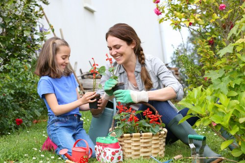 A woman and a young girl are sitting on a lush green lawn in a well-maintained garden, tending to potted plants with red flowers. The woman, wearing a grey plaid shirt and green gardening gloves, is smiling as she helps the girl, who is dressed in a blue t-shirt and jeans. Surrounding them are various garden elements including a wooden basket filled with more flowering plants, a small watering can, and a decorative container. The garden is bordered by dense greenery, including shrubs, flowering bushes, and a variety of plants with pink and purple flowers. In the background, there is a white exterior wall of a house with visible planter boxes, suggesting a tranquil outdoor space in Maida Vale, London. The scene is bathed in natural daylight, capturing an active moment of gardening and outdoor maintenance, illustrating services related to garden care and plant cultivation, with a focus on creating vibrant, peaceful garden environments typical of the local area.