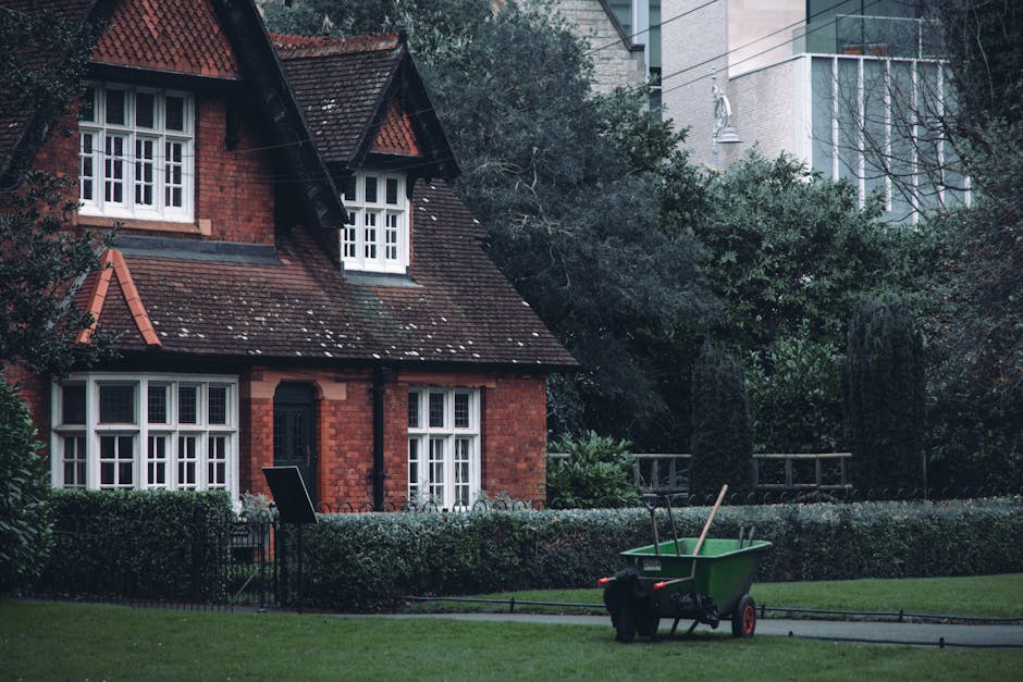A red-brick, two-story house with a steeply pitched tiled roof and white-framed windows is set amidst a well-maintained garden. The garden features a neatly trimmed hedge bordering a lush green lawn w