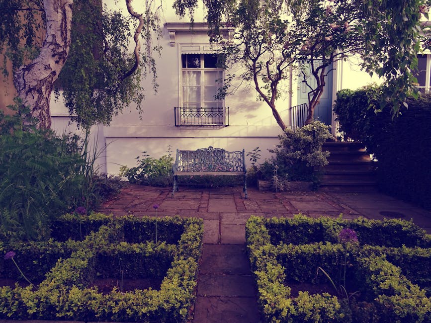 A red-brick, two-story house with a steeply pitched tiled roof and white-framed windows is set amidst a well-maintained garden. The garden features a neatly trimmed hedge bordering a lush green lawn w